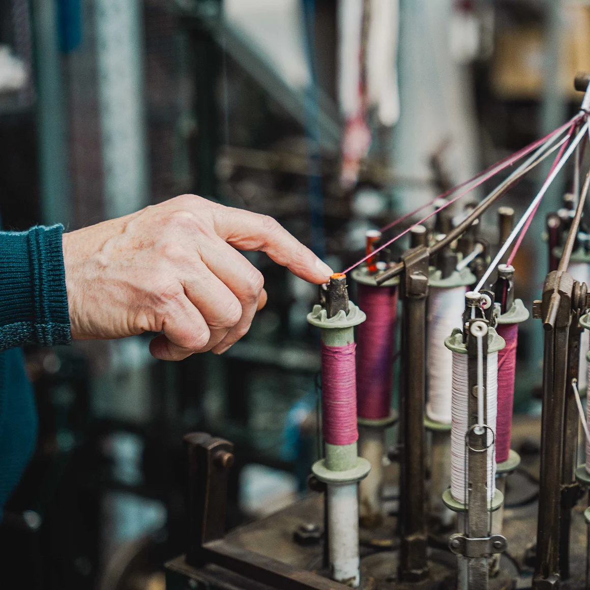 Hand wijst naar kleurrijke garenspoelen op een historische textielmachine in het Weverijmuseum.